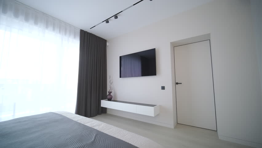 A contemporary minimalist bedroom interior, focusing on a large bed with a dark charcoal frame, crisp white and grey layered bedding, plump white pillows, and a textured throw blanket, set against light beige textured walls. Natural daylight illuminates the scene from a large floor-to-ceiling window on the left.