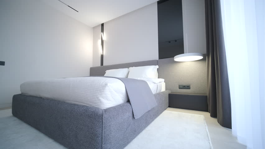 A contemporary minimalist bedroom interior, focusing on a large bed with a dark charcoal frame, crisp white and grey layered bedding, plump white pillows, and a textured throw blanket, set against light beige textured walls. Natural daylight illuminates the scene from a large floor-to-ceiling window on the left.
