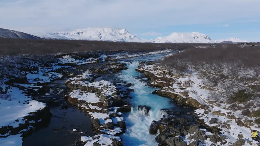Stunning aerial view of Bruarfoss waterfall in winter, Iceland. Turquoise blue water flows through snowy volcanic landscape with majestic snow-capped mountains in background.