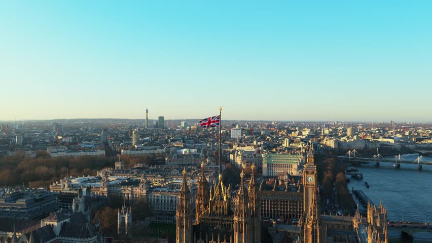 Waving Union Jack flag on top of Victoria Tower in London with Big Ben and River Thames while camera orbits the Houses of Parliament on a winter sunset