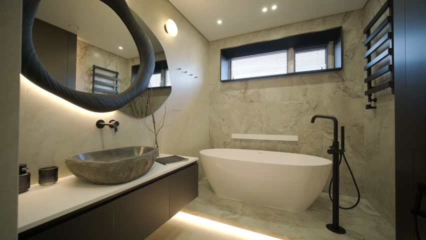 Luxury modern bathroom interior featuring a white freestanding bathtub and a unique stone vessel sink. The room is decorated with marble tiles, a large oval backlit mirror, and black matte fixtures in a minimalist contemporary style.