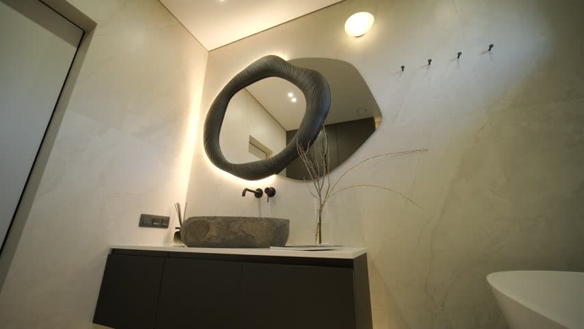 Luxury modern bathroom interior featuring a white freestanding bathtub and a unique stone vessel sink. The room is decorated with marble tiles, a large oval backlit mirror, and black matte fixtures in a minimalist contemporary style.