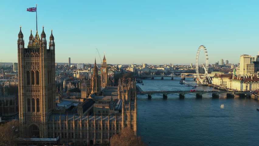 Aerial recording of Palace of Westminster with River Thames flowing nearby, showcasing iconic landmarks like London Eye and Big Ben under a clear sky on a bright, sunny day