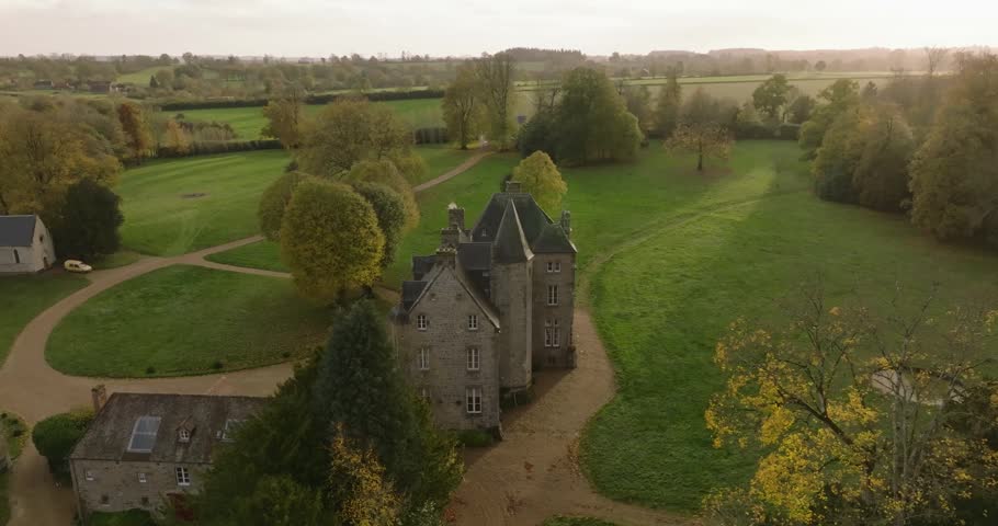 Stunning drone footage of historic French chateau with stone towers surrounded by autumn trees and rolling green countryside. Golden hour lighting creates warm atmospheric mood across the pastoral