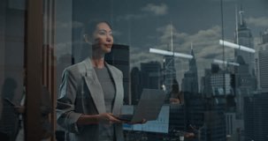 Asian Businesswoman Stands With a Laptop by an Office Window, Thinking Through Startup Growth Strategy as Her Leadership Focus Brings Success. Cinematic Reflections of Big City Financial District - Powered by Shutterstock - Get 15% off with code: PIKWIZARD15