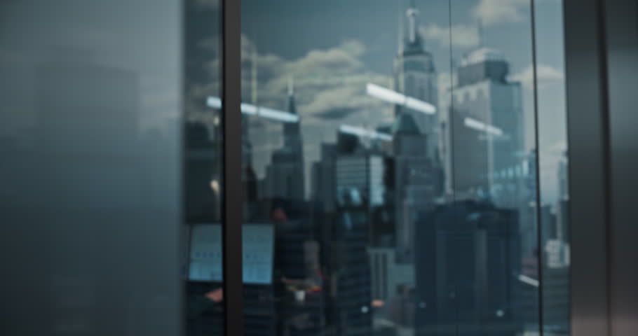 Young Caucasian Businessman in a Gray Suit Stands by the Office Window, Hand on Chin, Focused on Strategy as Reflections Reveal City Skyscrapers, Bright Workspaces, and Successful Colleagues in Motion
