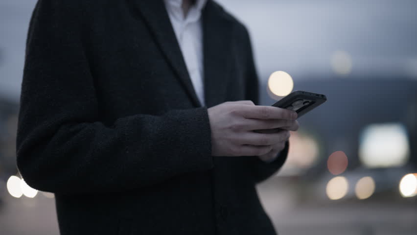 A young business professional stands on an urban street, focused on his smartphone. He is engaged in digital communication amidst the blurred city lights.