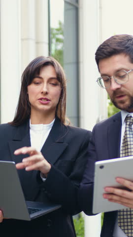 A focused businesswoman points at the laptop screen while walking through a city district with her successful male colleague, who is holding a tablet. They are discussing a new project together