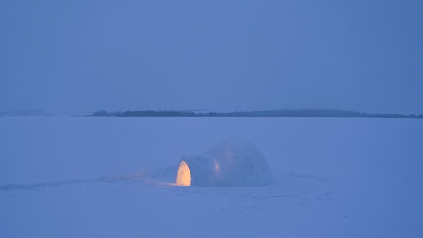 Lonely igloo glowing with warm light on frozen snowy field during snowfall. Winter night landscape, snow falling over arctic style shelter
