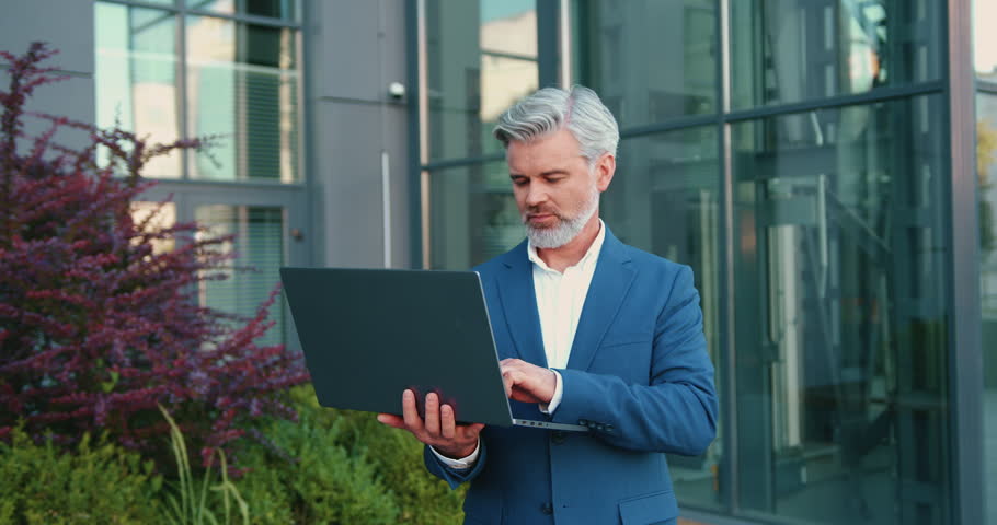 Portrait of a man entrepreneur carrying a laptop and looking at camere on city streets among modern corporate buildings. Business career and professional lifestyle concept.