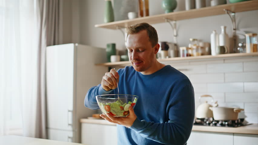 Hungry man enjoy salad sitting modern kitchen interior closeup. Happy male vegan eating tasty fresh vegetables meal closing eyes feeling satisfying. Smiling healthy guy chewing vitamin rich food home.