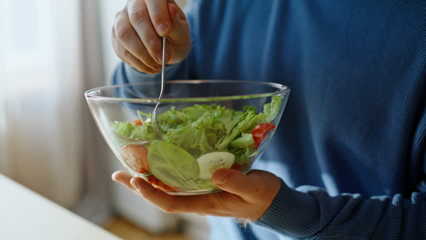 Closeup man eating salad from glass bowl enjoying fresh organic ingredients taste sitting apartment kitchen alone. Portrait smiling young guy chewing vitamin vegetables home. Vegetarian diet concept