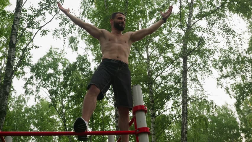 Shirtless male celebrating on top of red exercise frame, arms raised in triumphant pose, smiling against canopy of birch trees, playful acrobat vibe, crowdfree forest setting, energized