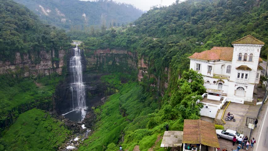 Drone pans from Tequendama waterfall to a white cliffside house, revealing lush green canyon walls, roadside terraces and small groups of tourists under misty skies.