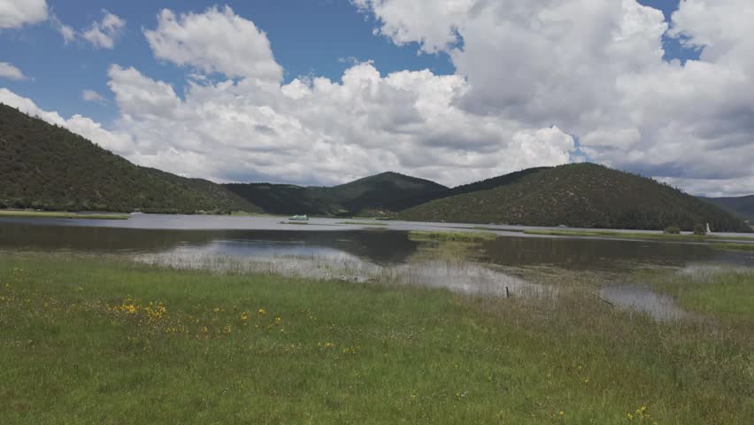 A breathtaking wide shot of Potatso National Park featuring the calm Shudu Lake surrounded by lush green meadows and rolling hills under a cloudy blue sky.
