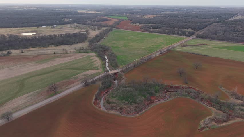 Aerial drone view of winding creek bend and rural bridge through red dirt farmland with green fields emerging in early spring Oklahoma countryside.