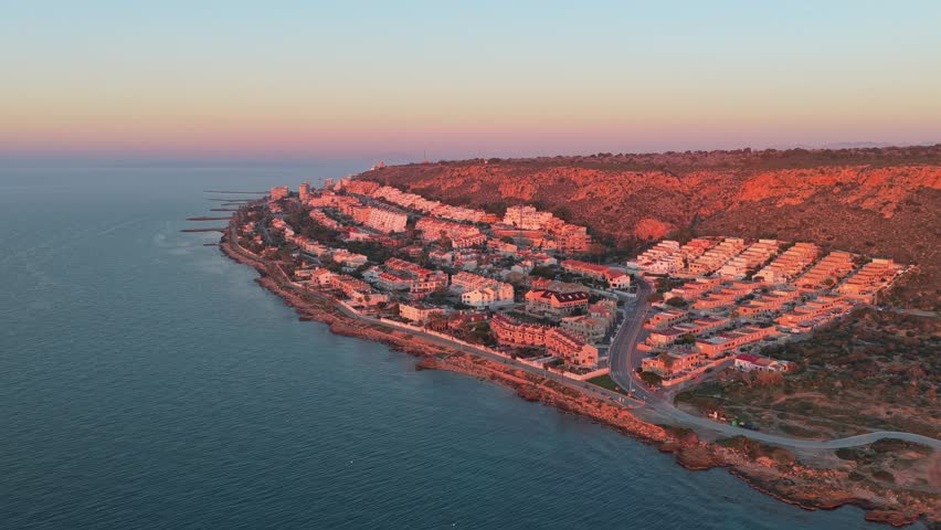 Stunning aerial footage capturing the beautiful coastal town of santa pola, spain, as the sun sets, casting a warm, golden glow over the buildings and sea