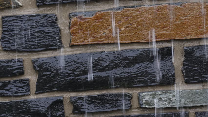 Raindrops fall vertically onto a brick wall. Bricks show black, brown, and gray tones. Water streaks down the textured surface. Close-up view captures rain