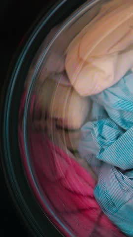Frontload washer spinning colorful clothes, rotating drum blur creates swirling textile motion, closeup through glass shows mix of stripes and bright towels, suds and water streaks catch light,