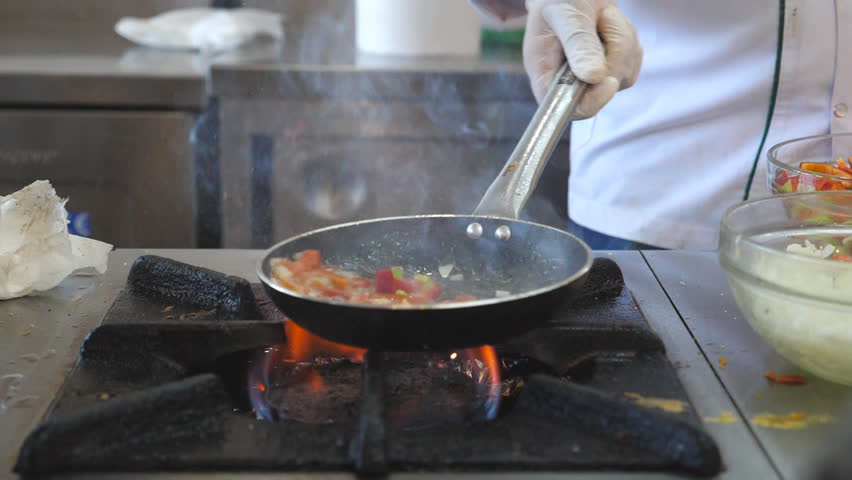 Unrecognizable chef cooking tasty flambe style dish on pan in modern kitchen of restaurant. Professional cook mixing and frying vegetables in flaming skillet on gas plate. Slow motion Front view