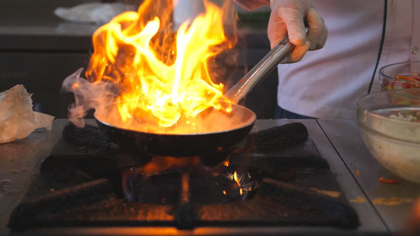 Unrecognizable chef cooking tasty flambe style dish on pan in modern kitchen of restaurant. Professional cook mixing and frying vegetables in flaming skillet on gas plate. Slow motion Front view