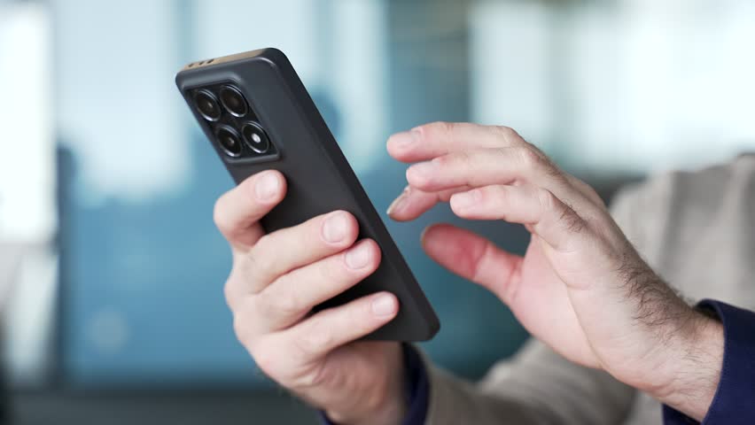 Close up of businessman hands using smartphone with blurred office background. Professional browsing mobile phone screen, typing message and checking online information on digital device at workplace.