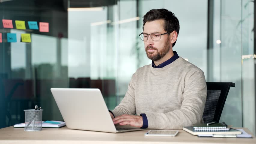 Confused businessman working on laptop at desk in office, then shrugging shoulders and spreading hands while looking at camera. Uncertain mature professional showing doubt and hesitation at workplace.