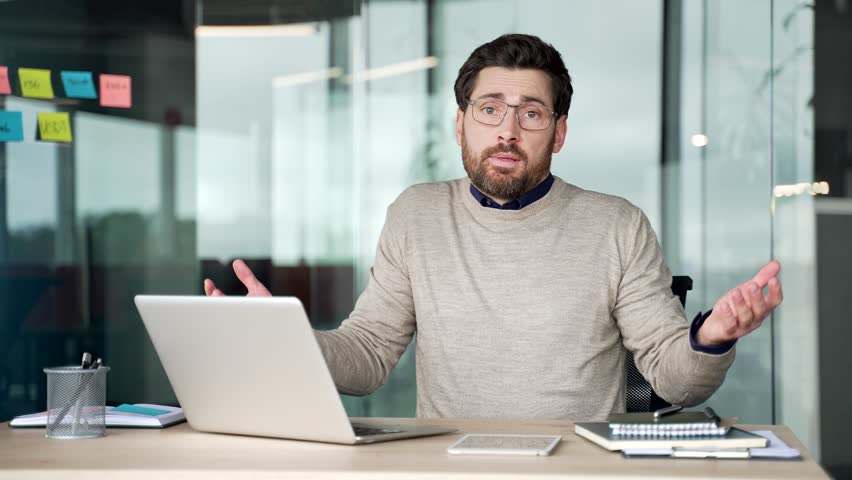Confused businessman working on laptop at desk in office, then shrugging shoulders and spreading hands while looking at camera. Uncertain mature professional showing doubt and hesitation at workplace.