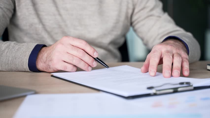 Close up of businessman hand signing documents with pen while sitting at desk in modern office. Executive CEO in casual sweater approves contract, completing agreement and business paperwork.
