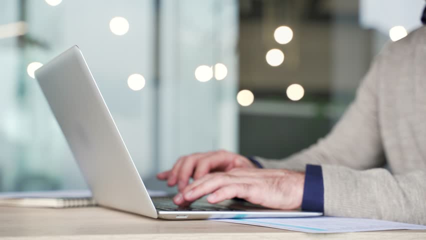 Close up of businessman hands typing on laptop keyboard at desk in modern office. Mature professional working on computer, entering text, managing information during daily business tasks at workplace.