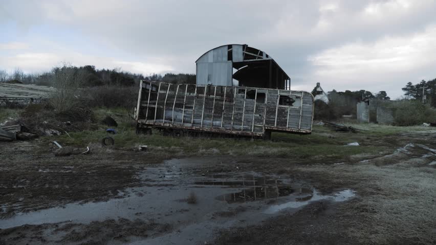 Abandoned Farm scene showcases the skeletal frame of a dilapidated structure and an old trailer sitting in a muddy field under moody grey clouds depicting rural decay and forgotten history