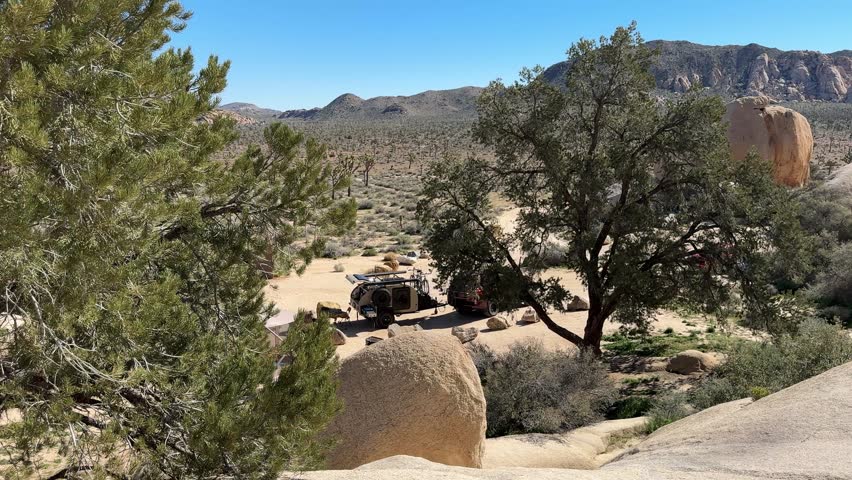 Teardrop camper parks among large rocks in Joshua Tree National Park under clear skies in California during daytime.