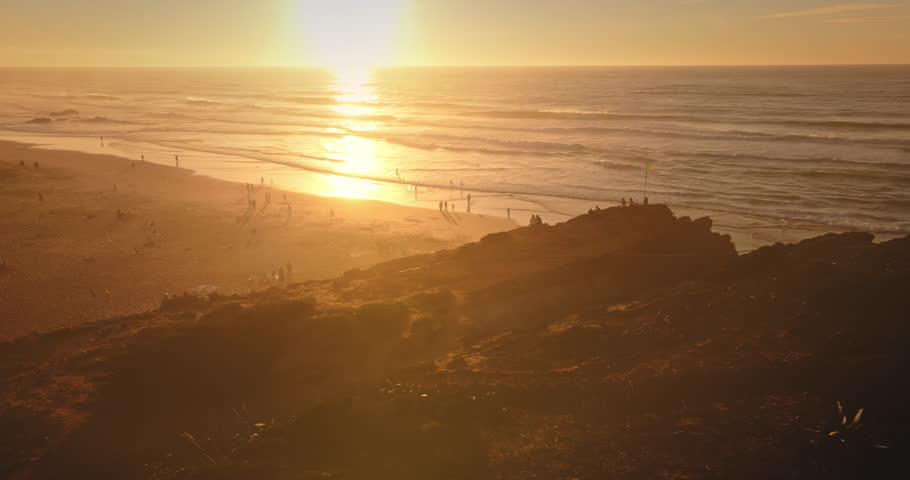 Golden sunlight bathing Praia do Guincho beach and Atlantic ocean waves during a vibrant sunset, people enjoying the sandy shore and natural park landscape. Summer vacation. Aerial flight panorama