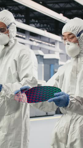 Vertical video Multiracial team examining semiconductor substrate in cleanroom production plant. Diverse coworkers wearing protective gear verify optical photolithography patterns on silicon wafers