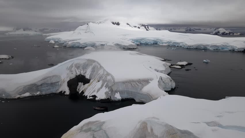 Cinematic aerial flying forward from one ice shelf to another over the dark waters of Foyn Harbour, revealing the schooner Amazone moored between the massive blue ice walls of Antarctica
