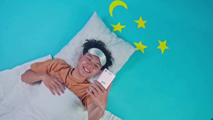 Top view of smiling man relaxing in bed holding mobile phone and gesturing thumb up on blue studio set