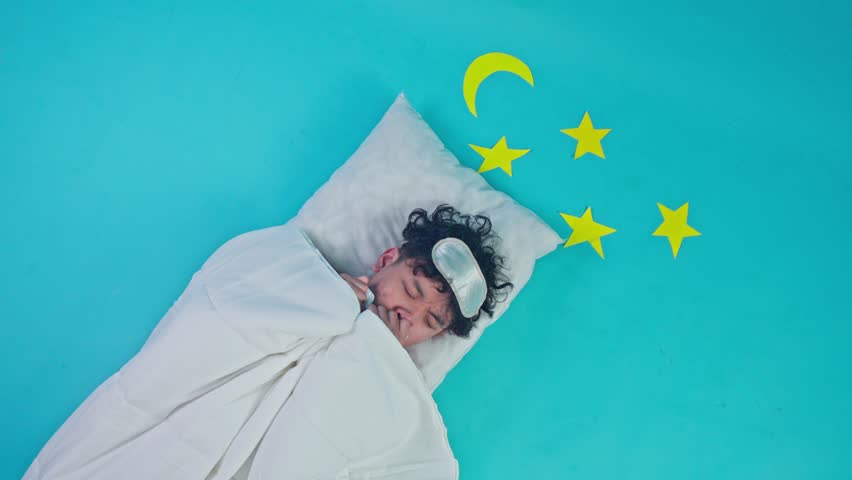 Top view of young man with sleep mask having nightmare or shivering under white blanket on blue background