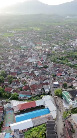 Aerial view of a small town with dense housing, roads, and rice fields, surrounded by green landscapes and mountains in the background