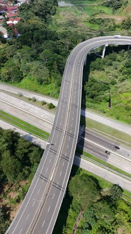 Aerial view of a curved highway overpass crossing a main road, surrounded by dense greenery and hillside terrain under bright daylight.