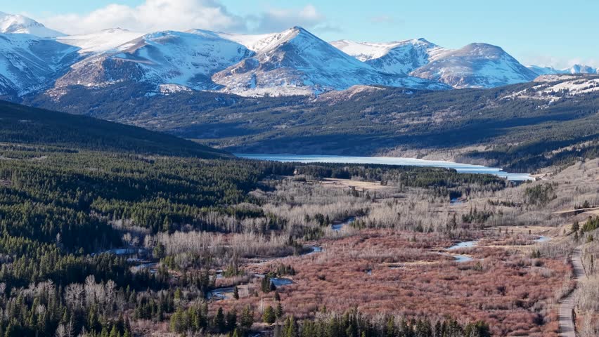 Wide aerial shot of a valley in Montana with snow-capped mountains towering above, evergreen forests, and rolling hills under a clear sky.