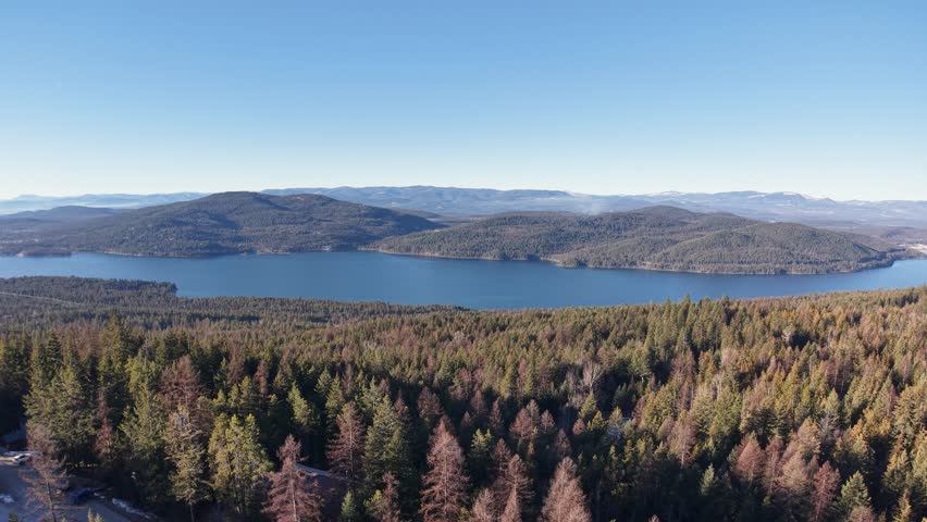 Aerial drone view of a vast mountain lake in Montana, with evergreen forests, a winding road, and hills across the water under a clear blue sky.