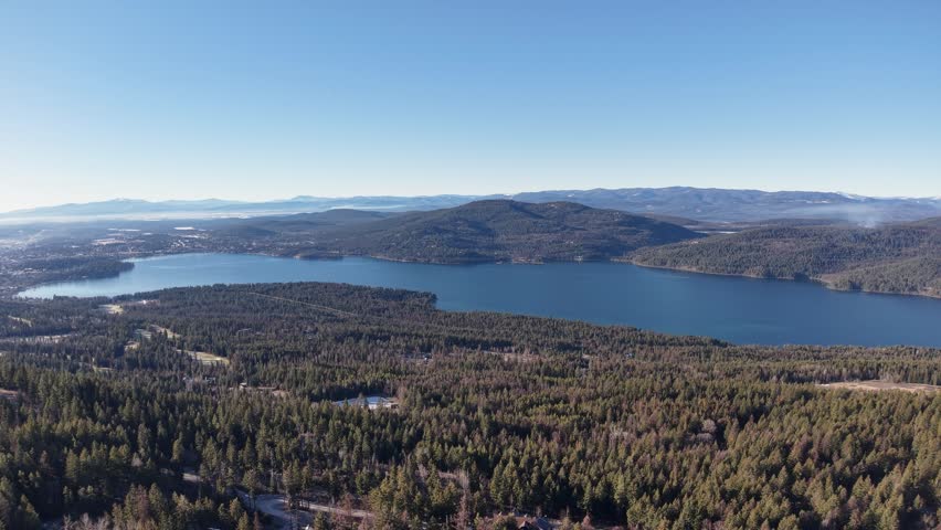 Aerial view of a tranquil mountain lake in Montana surrounded by dense evergreen forests, with rolling hills and mountains in the distance beneath a clear blue sky.