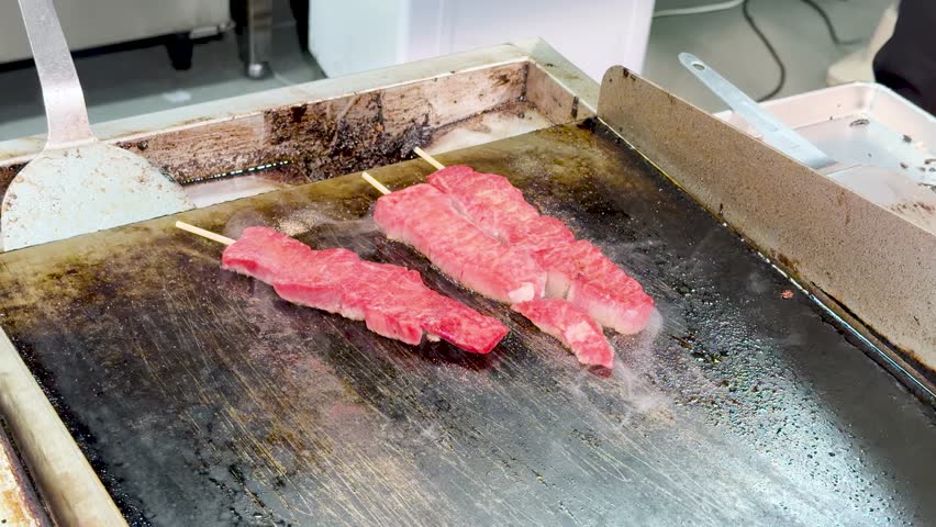 Raw marbled beef skewers sizzle on a flat metal grill at a Japanese street market