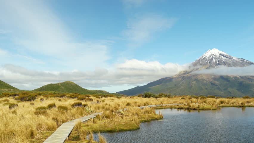Panning landscape shot at Pouakai Tarns in New Zealand, revealing the reflective wetland, golden tussock and Mount Taranaki rising beyond under low drifting cloud