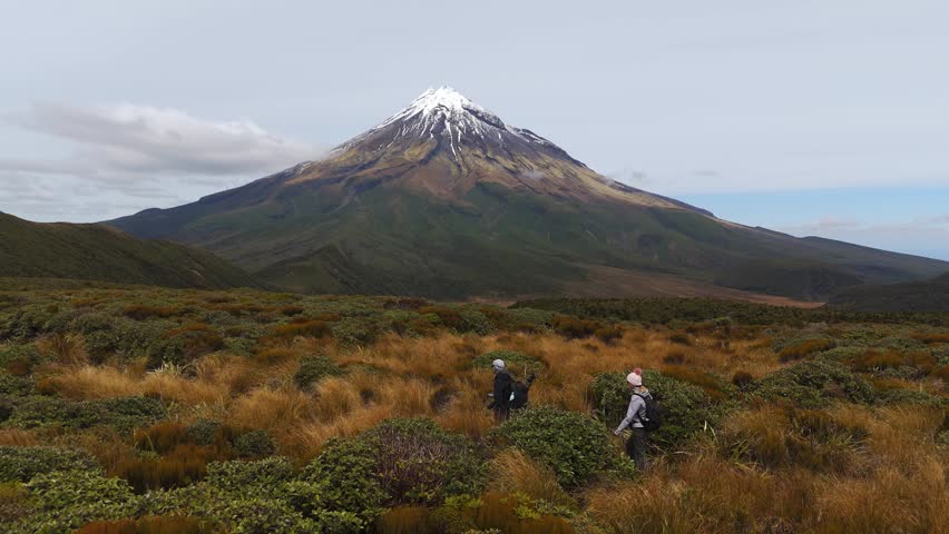 Young couple hiking across alpine scrub with Mount Taranaki in New Zealand rising behind under soft cloud and open sky, cinematic tracking aerial shot
