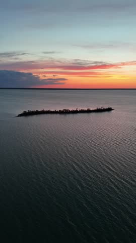 Vertical drone push-in toward Goat Island at Steep Rock Manitoba over calm Lake Winnipeg during summer sunset, showing orange-red skies, shoreline, and surrounding trees.