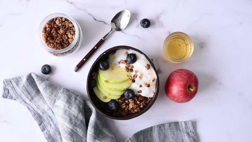 Natural greek yogurt with granola, blueberry and apple in a bowl on a marble background with honey, fruit, berries, napkin and spoon. Healthy and nutritious breakfast concept. Top view, copy space.