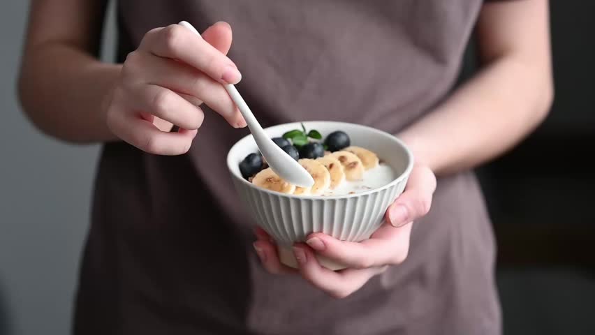 Woman holding bowl of granola with greek yogurt, banana, blueberries and spoon in hands on a brown t-shirt background close up. Milk breakfast concept, oat muesli in natural light. Front view, copy space.