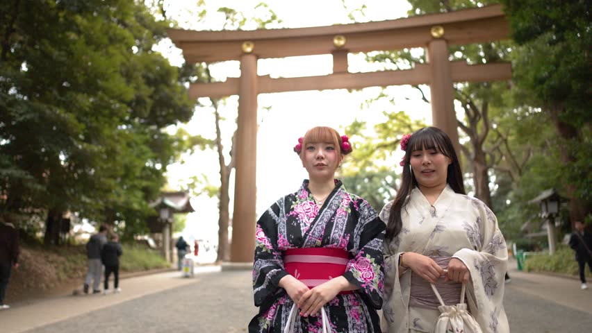 Two women wearing traditional kimonos stand smiling before a large wooden torii gate in Tokyo