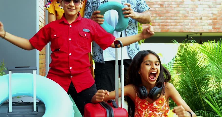 Cheerful Indian family of four in vacation attire posing on a home porch with joy. Happy parents and kids with suitcases and bags ready for a summer holiday trip